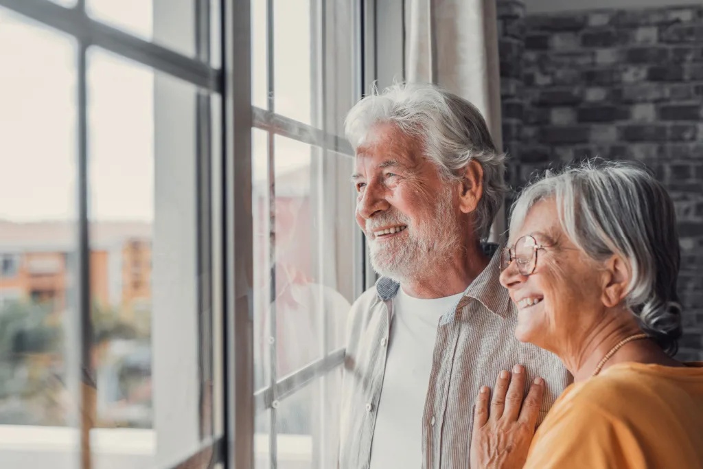 older man and woman smile while looking out a window onto their neighborhood street