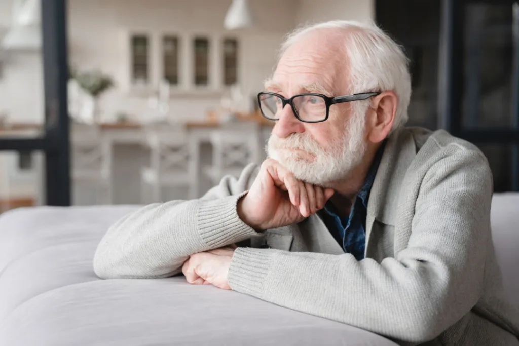 elderly man wearing glasses leans over the backrest of a sofa to look out the window longingly