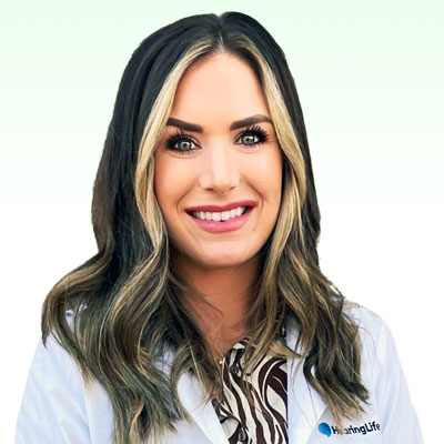 A woman with shoulder-length hair smiles, standing against a pale green background. She wears a white coat with