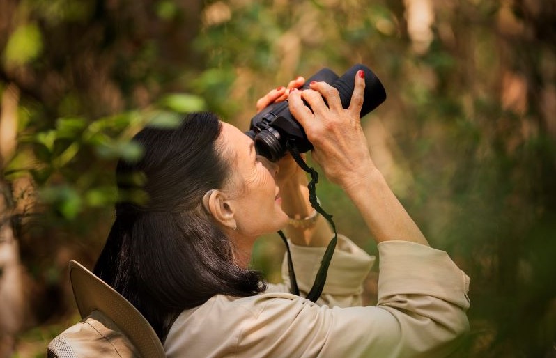 A woman wears hearing aids while looking through binoculars and birding. 